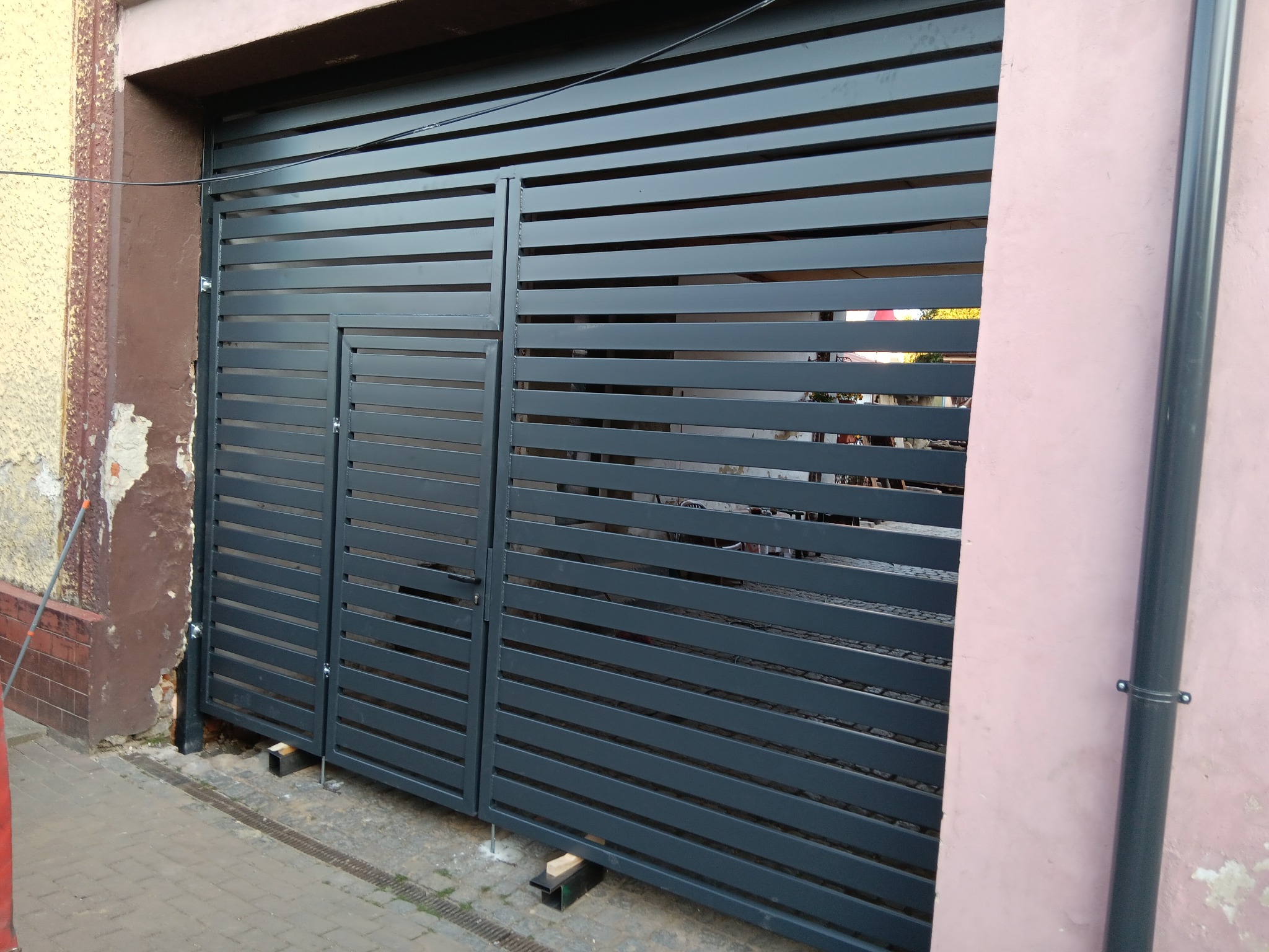 Nowoczesna, pozioma brama wjazdowa z metalowych profili, pomalowana na szaro, zamontowana między budynkami o zróżnicowanej elewacji. Widoczne detale mocowania.