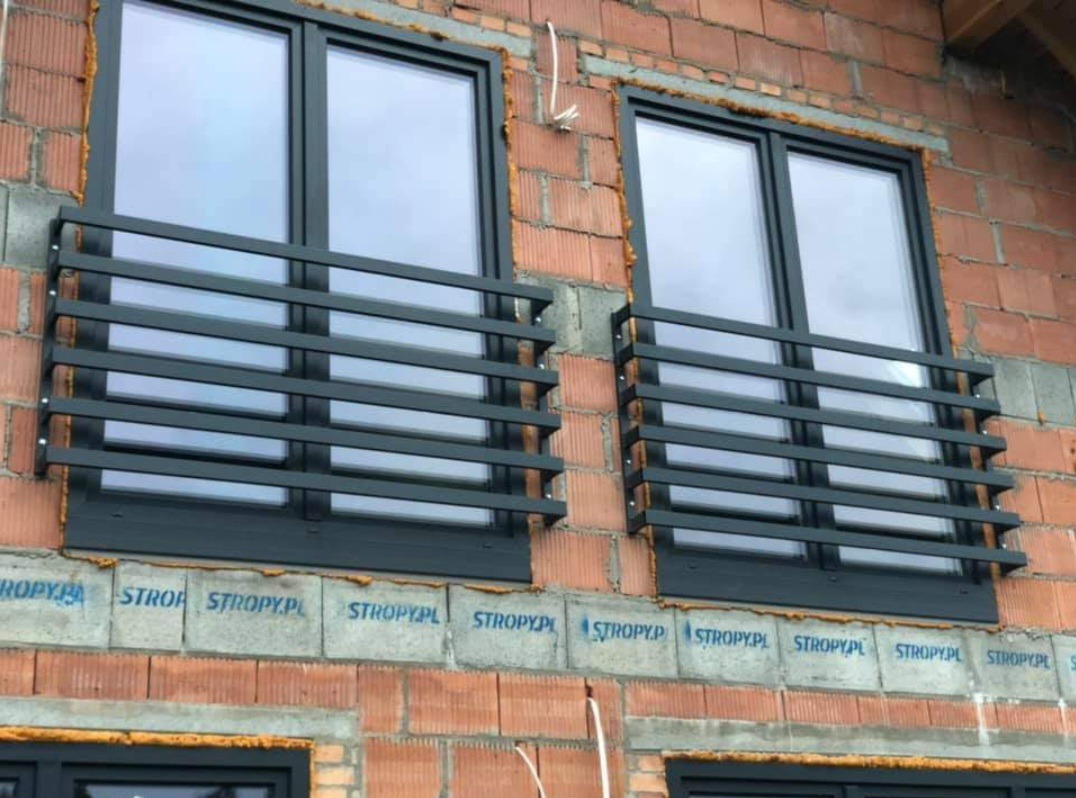 Dwa okna z czarnymi ramami i metalowymi balustradami ochronnymi na surowej ceglanej ścianie budynku w trakcie budowy. Widoczne detale montażu.