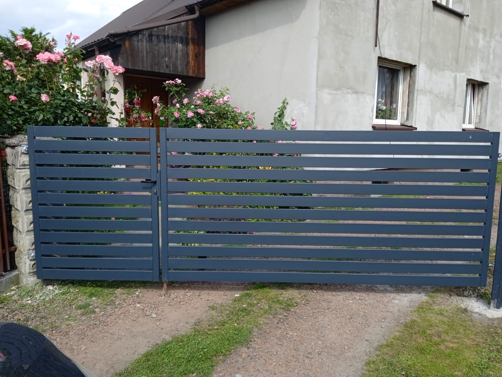 Nowoczesna, pozioma brama wjazdowa w kolorze grafitowym, z widocznymi różowymi kwiatami w tle. Brama zamontowana przed szarym domem.