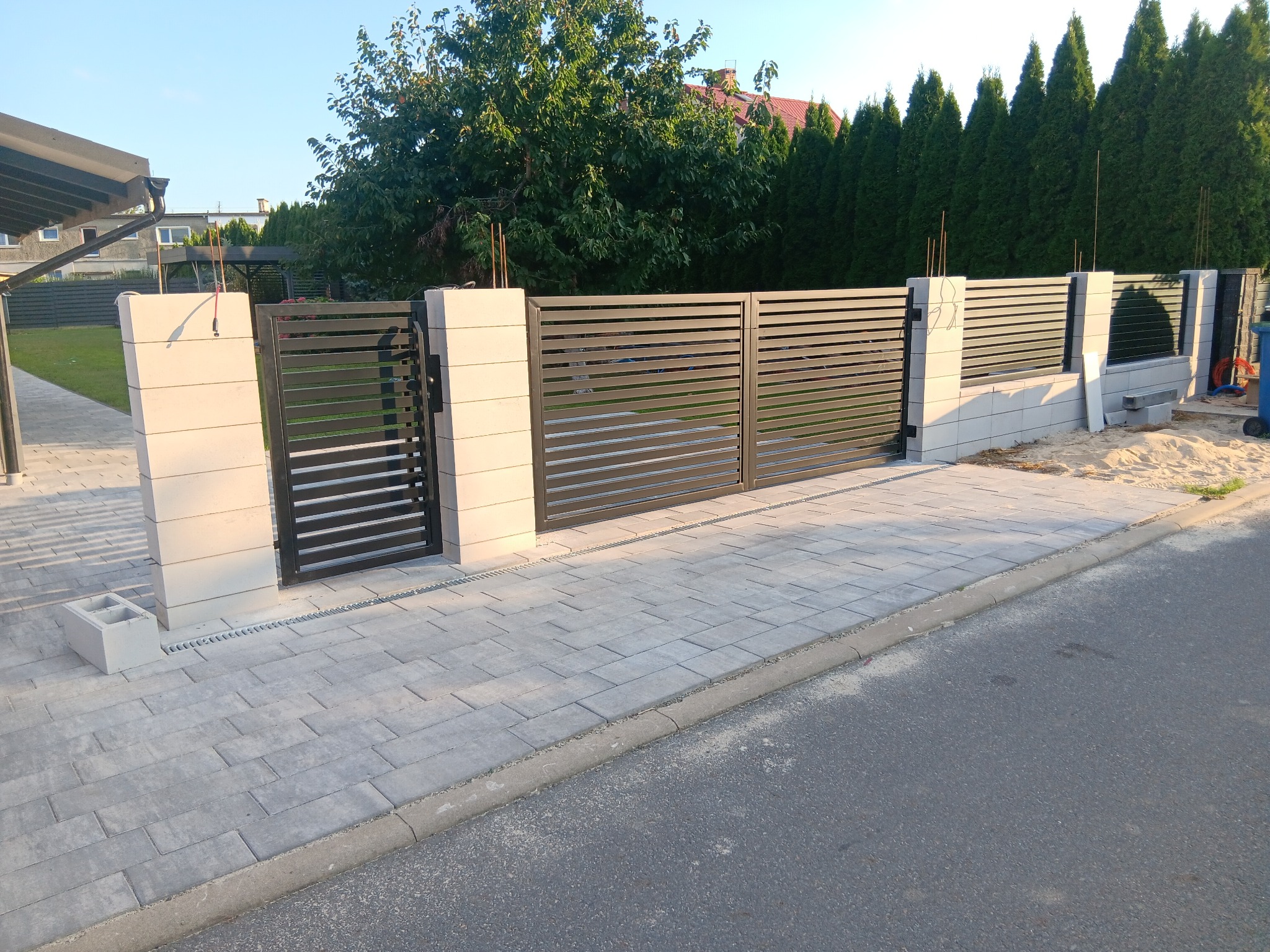 Nowoczesna brama wjazdowa z poziomymi panelami, osadzona na betonowych słupkach. Kostka brukowa prowadzi do asfaltowej drogi. W tle zieleń drzew i krzewów.