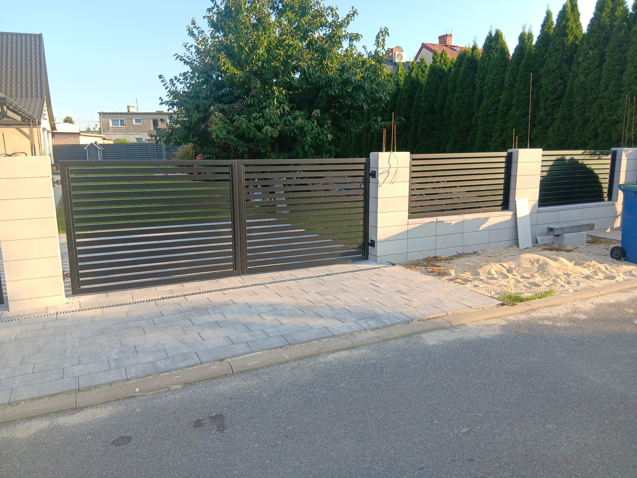 Nowoczesna, pozioma brama wjazdowa z metalowymi lamelami w kolorze grafitowym, osadzona na jasnych, betonowych słupkach. Widoczne kostki brukowe i zieleń w tle.