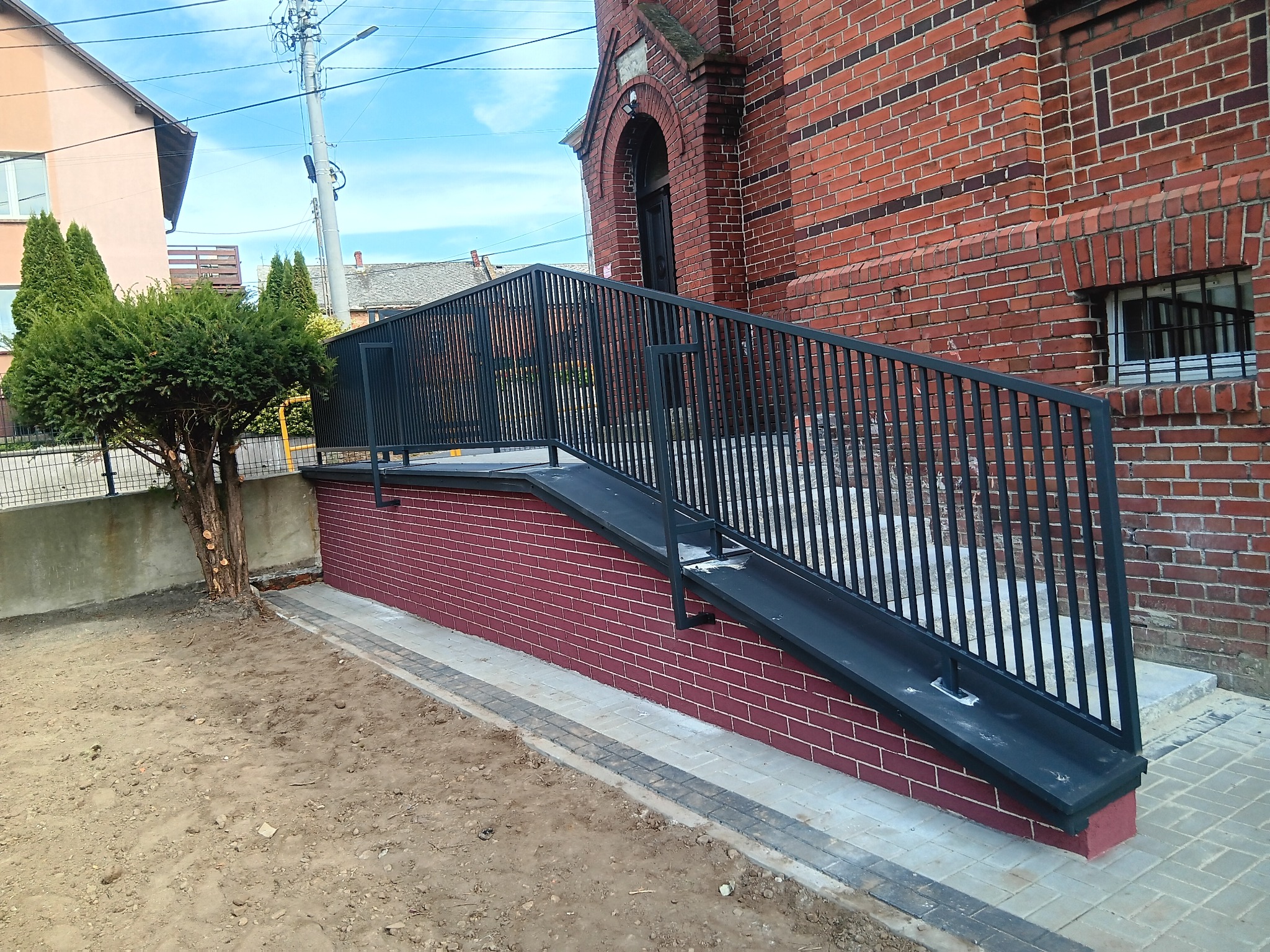 Rampa dla niepełnosprawnych z czarną, metalową balustradą przy ceglanym budynku. Widoczny fragment chodnika i rabata z drzewem. Słoneczny dzień, błękitne niebo.