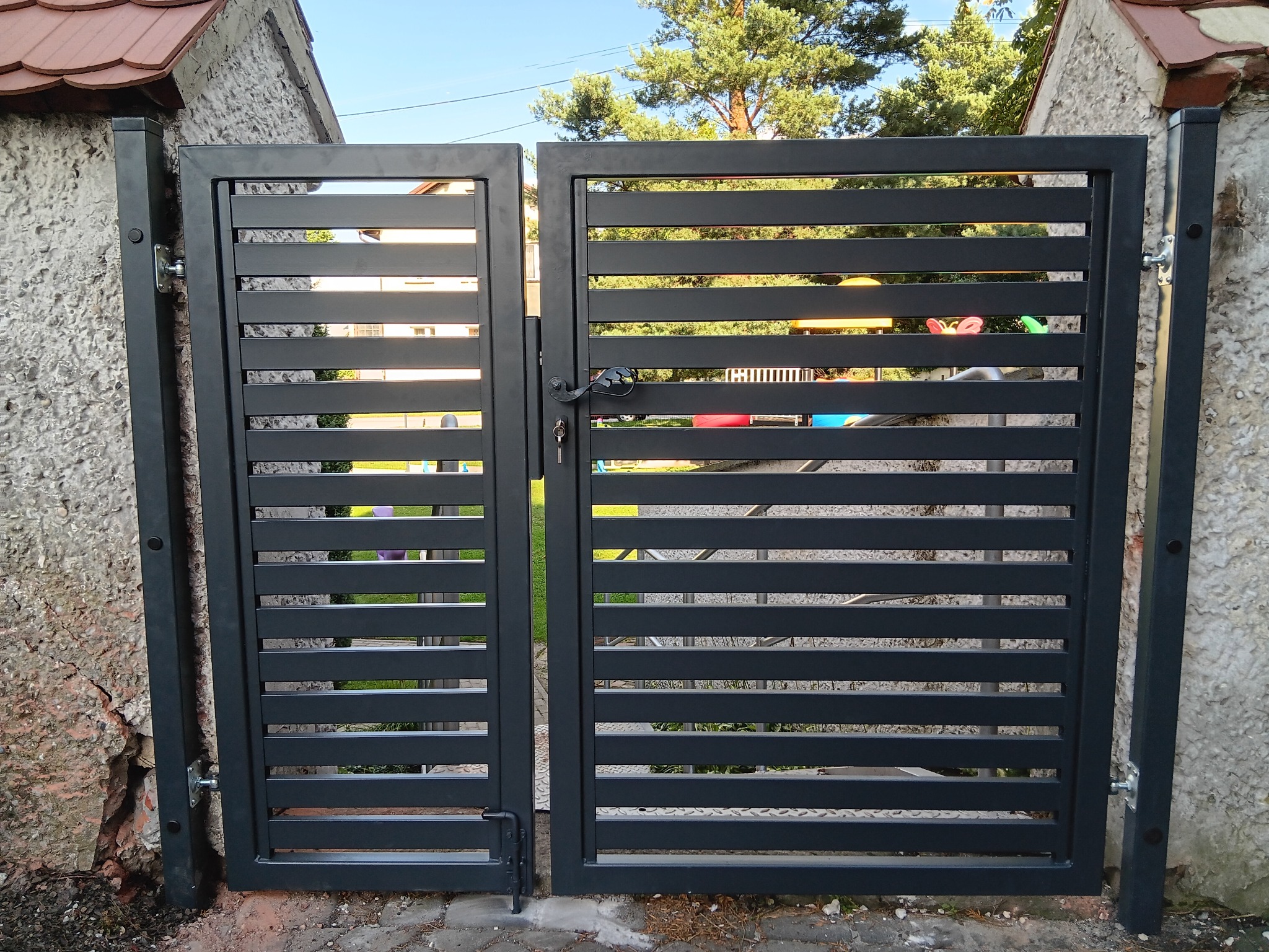 Nowoczesna, grafitowa brama wjazdowa z poziomymi szczeblami, zamontowana między budynkami o nierównych ścianach. Widoczny zamek i klucz. W tle plac zabaw.