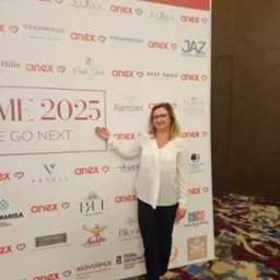 Kobieta w białej bluzce i czarnych spodniach stoi przed ścianką z logo hoteli i firm turystycznych, wskazując na napis 'ME 2025 WE GO NEXT'.