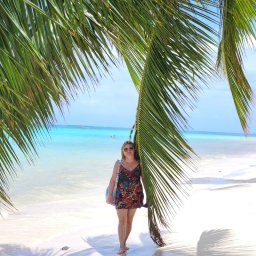 SUNNY4YOU SPÓŁKA Z OGRANICZONĄ ODPOWIEDZIALNOŚCIĄ - Kobieta w sukience spaceruje po białej, piaszczystej plaży w cieniu palmy, z turkusową wodą i błękitnym niebem w tle. Widok na egzotyczne wakacje.