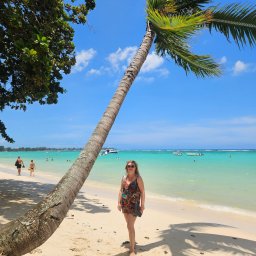 SUNNY4YOU SPÓŁKA Z OGRANICZONĄ ODPOWIEDZIALNOŚCIĄ - Kobieta w letniej sukience stoi na jasnej plaży pod palmą, z turkusową wodą i łodziami w tle, w słoneczny dzień. Widok na tropikalny raj.