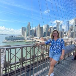 SUNNY4YOU SPÓŁKA Z OGRANICZONĄ ODPOWIEDZIALNOŚCIĄ - Uśmiechnięta kobieta w niebieskiej koszuli i dżinsowych szortach pozuje na tle panoramy Manhattanu z mostu Brooklińskiego w słoneczny dzień.