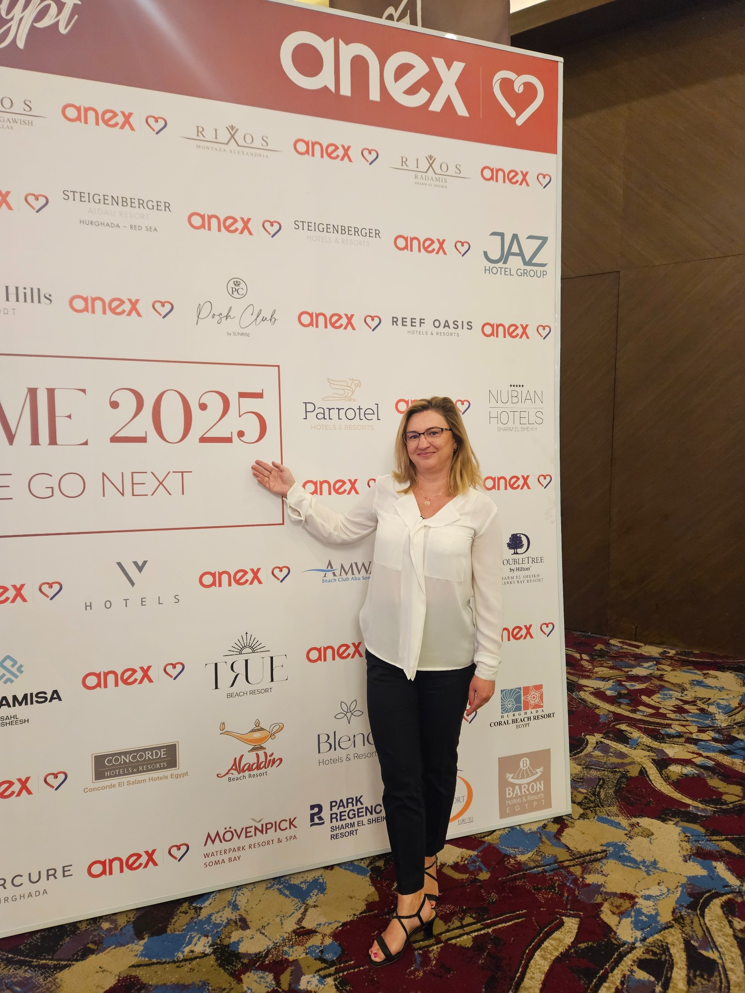 Kobieta w białej bluzce i czarnych spodniach stoi przed ścianką z logo hoteli i firm turystycznych, wskazując na napis 'ME 2025 WE GO NEXT'.