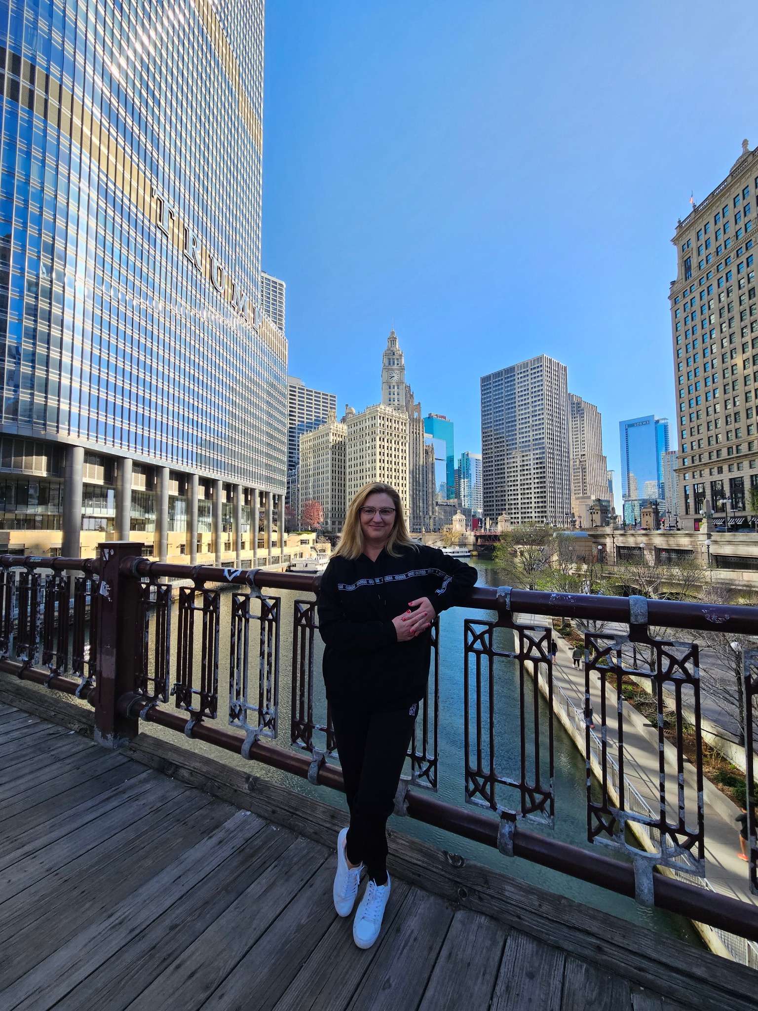 Kobieta w czarnym stroju opiera się o barierkę mostu na tle panoramy Chicago. W tle rzeka i drapacze chmur w słoneczny dzień.