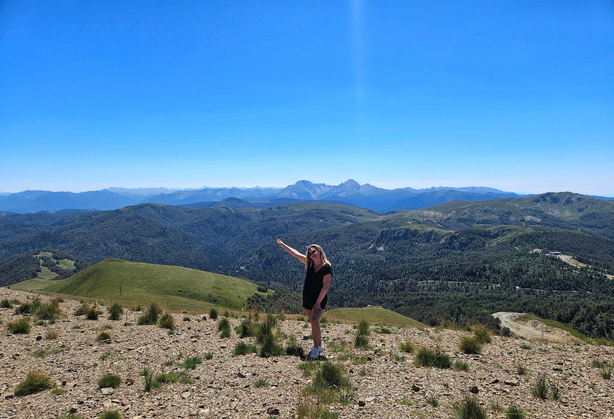 Kobieta w czarnej sukience na szczycie góry z rozległym widokiem na zalesione wzgórza i odległe pasmo górskie pod błękitnym niebem. Wyprawa górska.