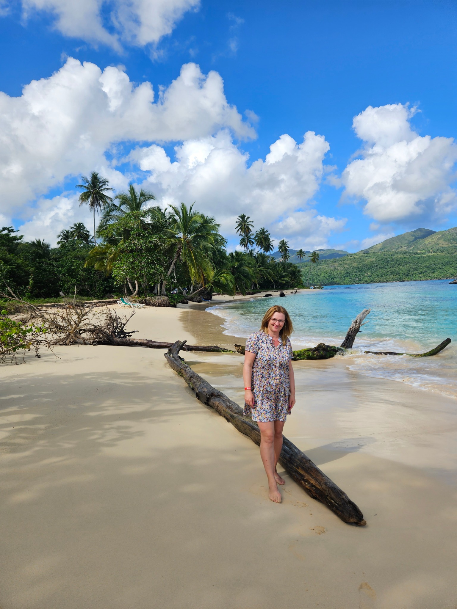 Kobieta w kwiecistej sukience stoi boso na piaszczystej plaży z powalonym pniem drzewa, w tle turkusowa woda, palmy i góry pod błękitnym niebem z chmurami.