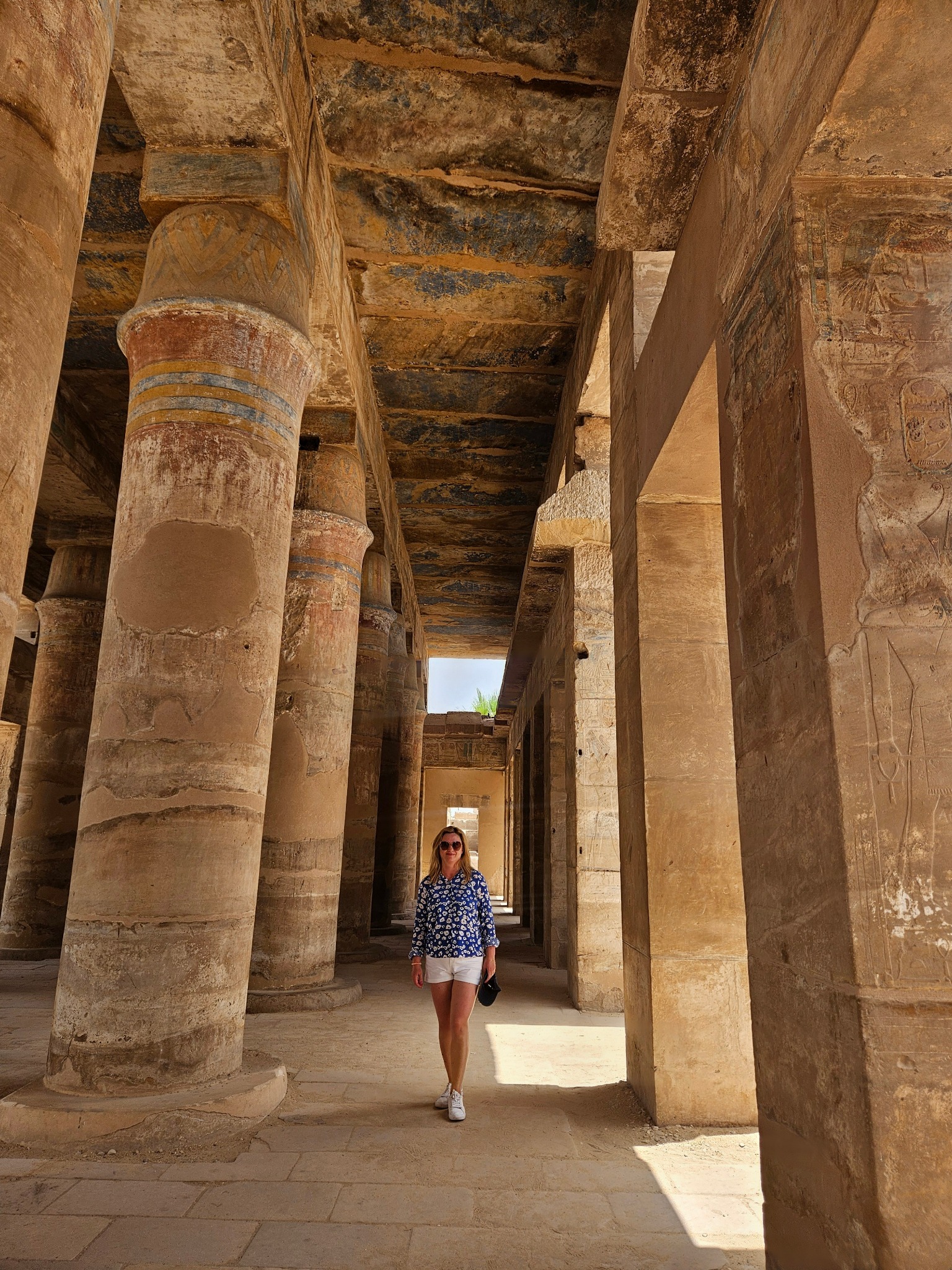 Kobieta w kwiecistej bluzce spaceruje po starożytnej świątyni z kolumnami. Światło słoneczne podkreśla detale architektoniczne i hieroglify na ścianach.