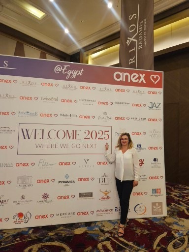 Kobieta pozuje przed ścianką z logo hoteli i napisem 'Welcome 2025 Where We Go Next' podczas wydarzenia branży turystycznej w Egipcie, pod banerem Rixos Radamis.