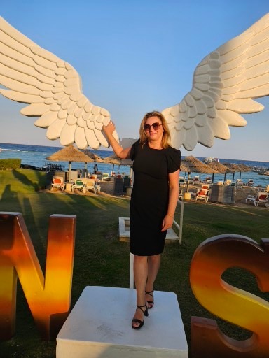 Kobieta w czarnej sukience pozuje na tle dekoracji z białymi skrzydłami anioła i napisu na plaży z leżakami i morzem w tle, prawdopodobnie podczas imprezy tematycznej.