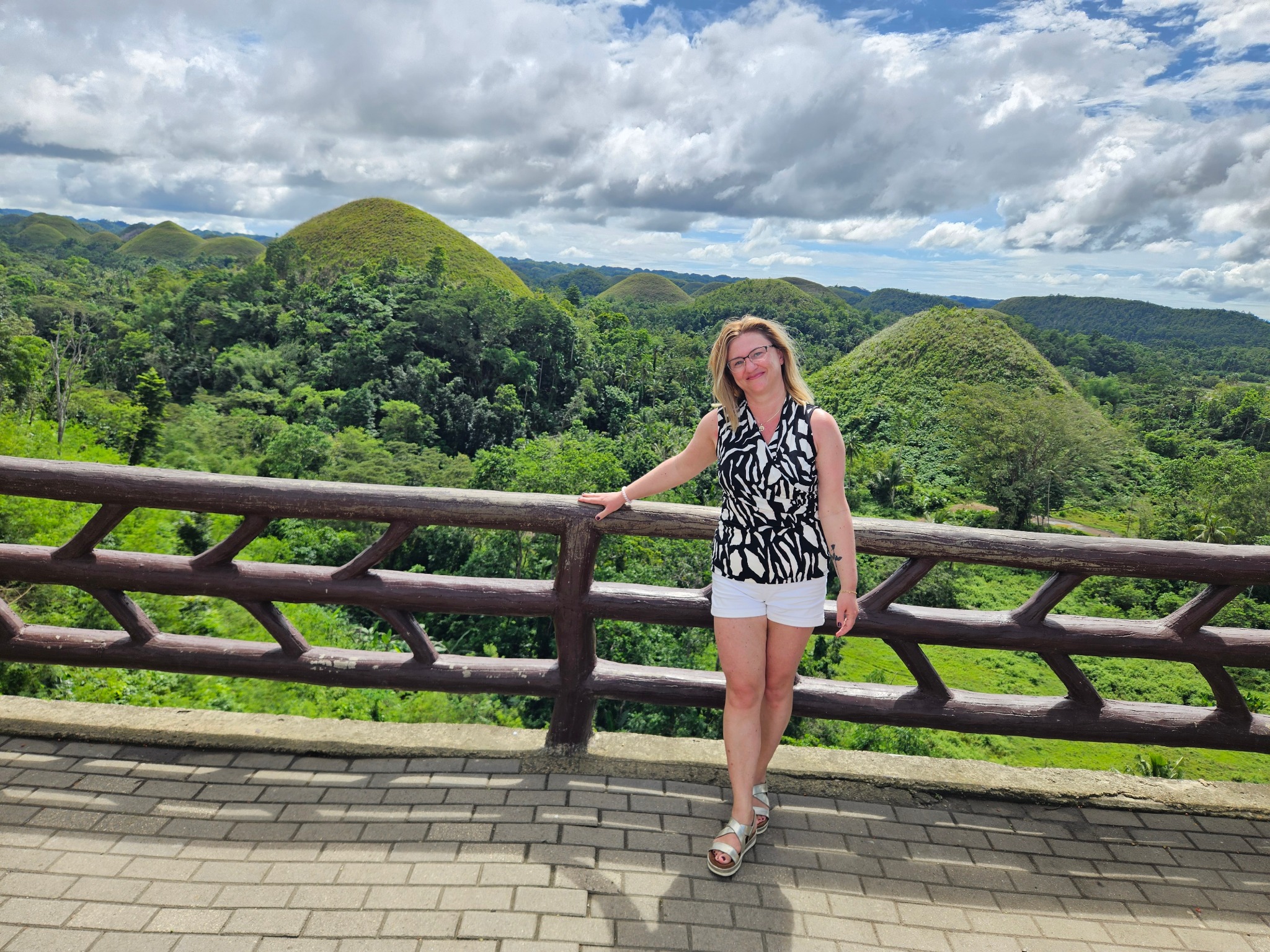Turystka na tle Czekoladowych Wzgórz na Filipinach. Kobieta w białych szortach i bluzce w zebrę opiera się o drewnianą barierkę, podziwiając malowniczy krajobraz.