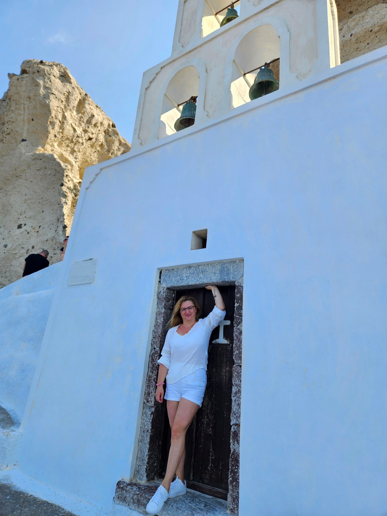 Kobieta w białym stroju pozuje w drzwiach białego kościoła z dzwonami na Santorini. W tle skała i fragmenty innych osób. Jasne, słoneczne światło.