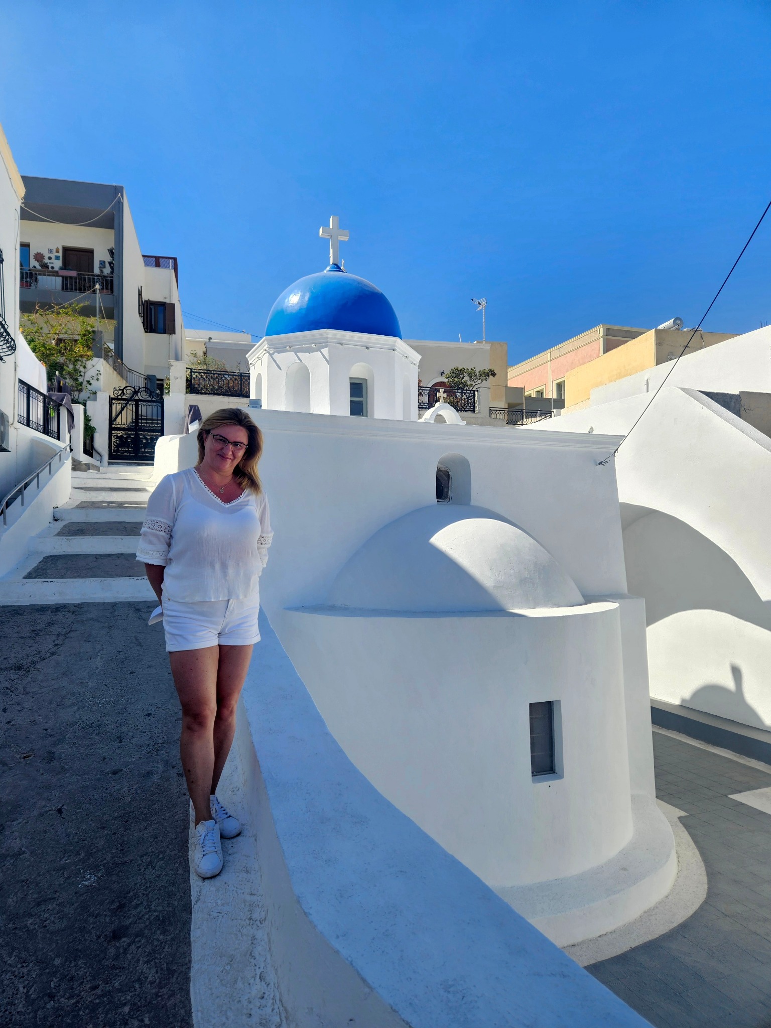 Kobieta w białym stroju pozuje na tle tradycyjnej greckiej architektury z białą cerkwią i błękitną kopułą na Santorini, w słoneczny dzień.