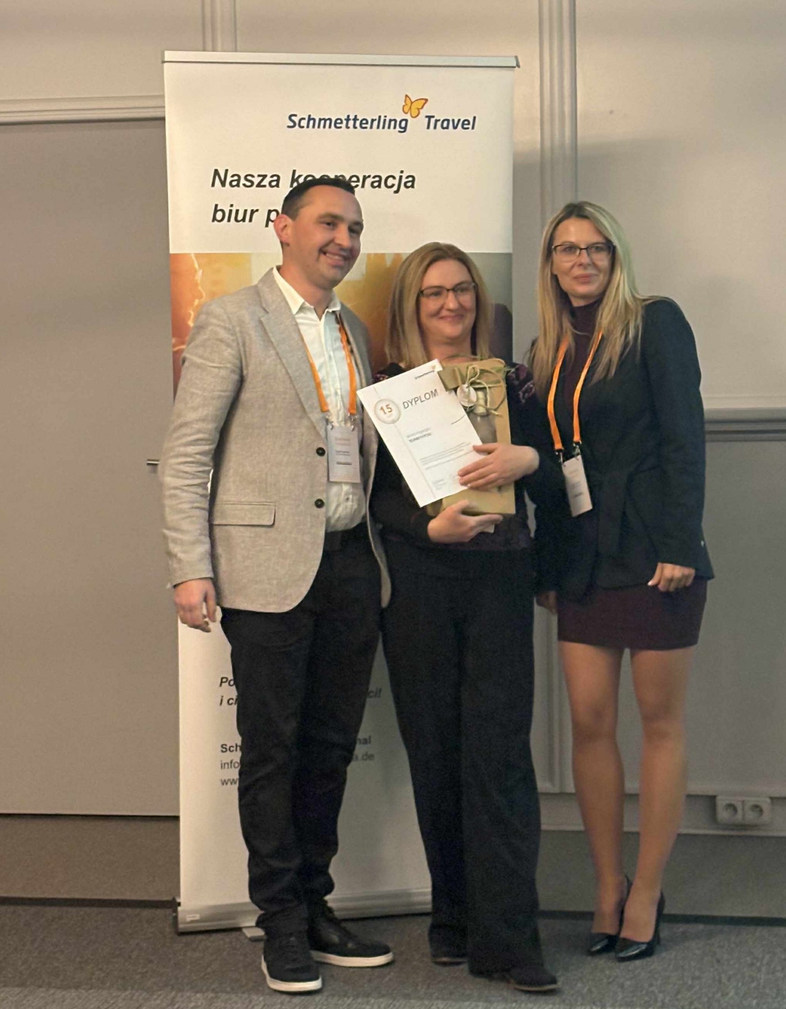 Wręczenie dyplomu dla Schmetterling Travel. Trzy osoby pozują z dyplomem na tle roll-upu z logo firmy. Uroczysta atmosfera, biznesowy charakter.