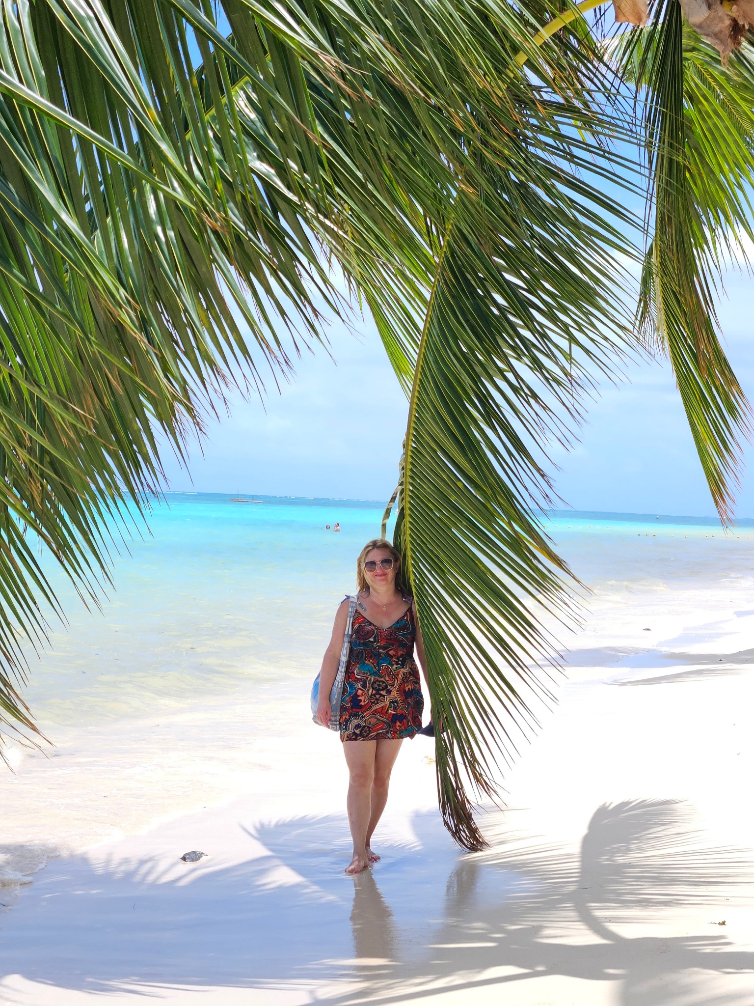 Kobieta w sukience spaceruje po białej, piaszczystej plaży w cieniu palmy, z turkusową wodą i błękitnym niebem w tle. Widok na egzotyczne wakacje.