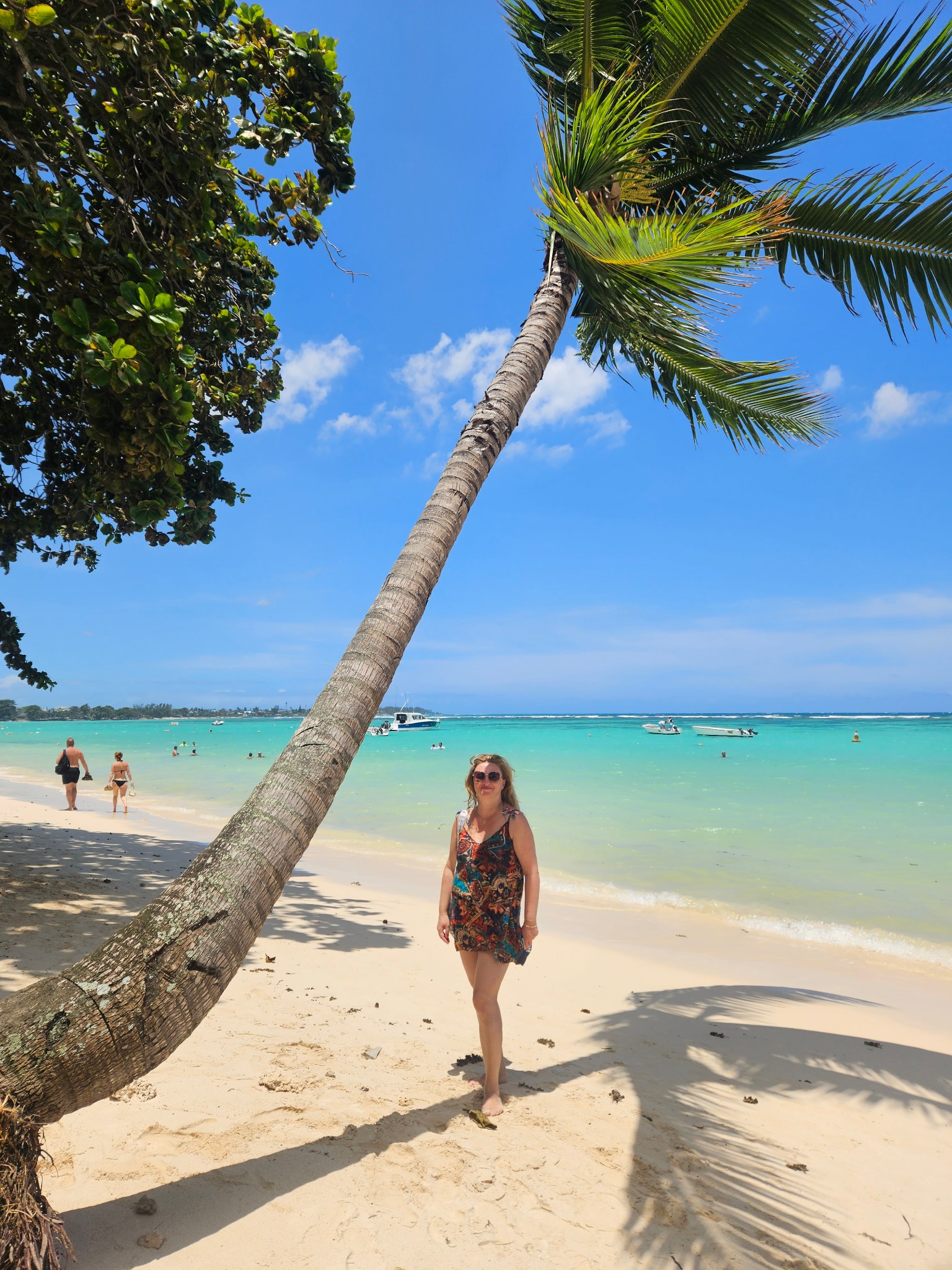 Kobieta w letniej sukience stoi na jasnej plaży pod palmą, z turkusową wodą i łodziami w tle, w słoneczny dzień. Widok na tropikalny raj.