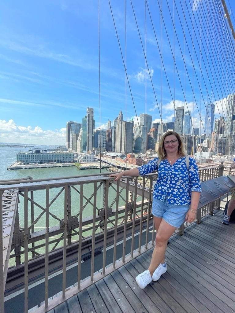 Uśmiechnięta kobieta w niebieskiej koszuli i dżinsowych szortach pozuje na tle panoramy Manhattanu z mostu Brooklińskiego w słoneczny dzień.