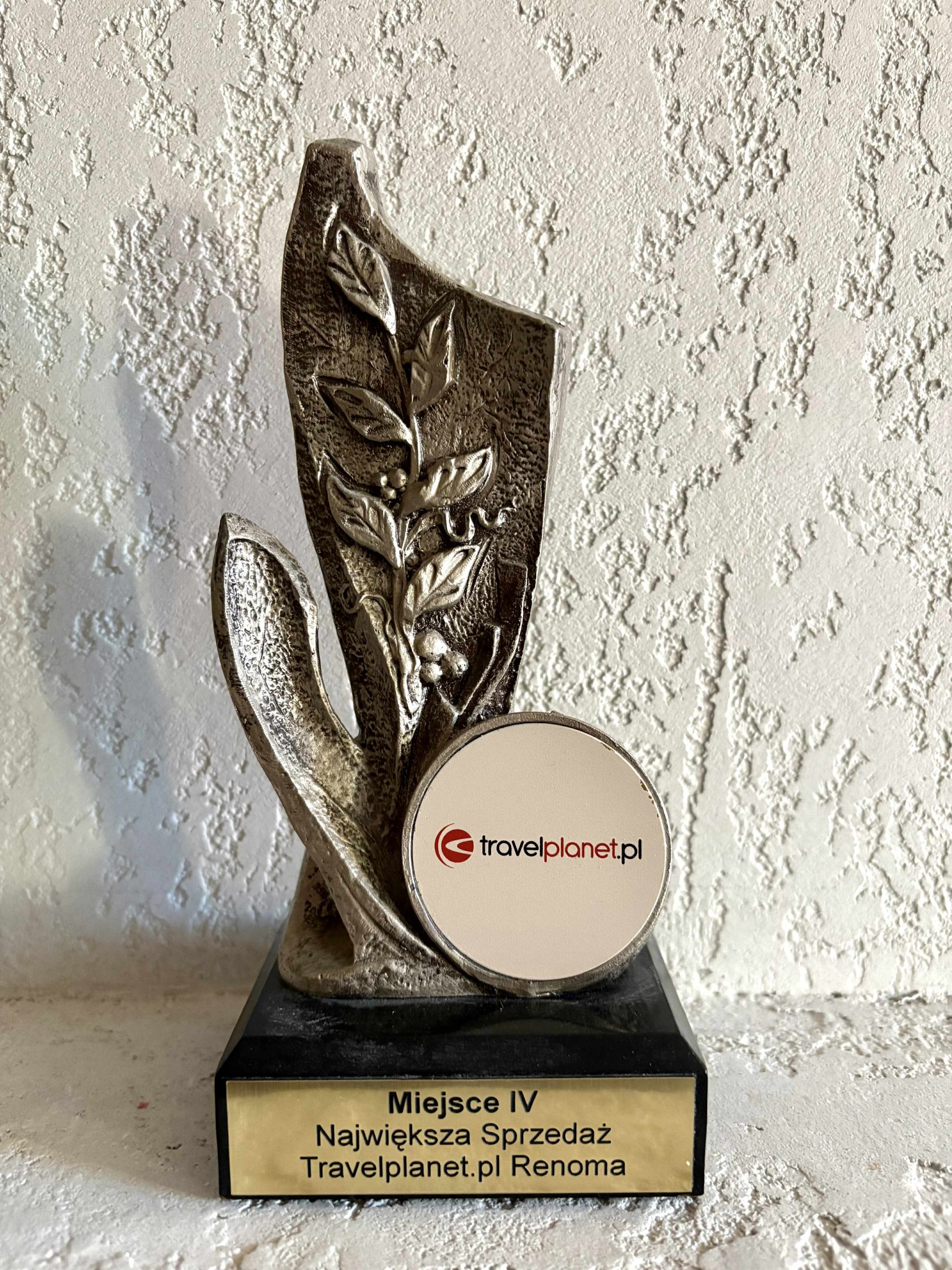 Statuetka Travelplanet.pl Renoma za IV miejsce w sprzedaży, z logo firmy na okrągłej naklejce, na tle białej ściany o nierównej fakturze.