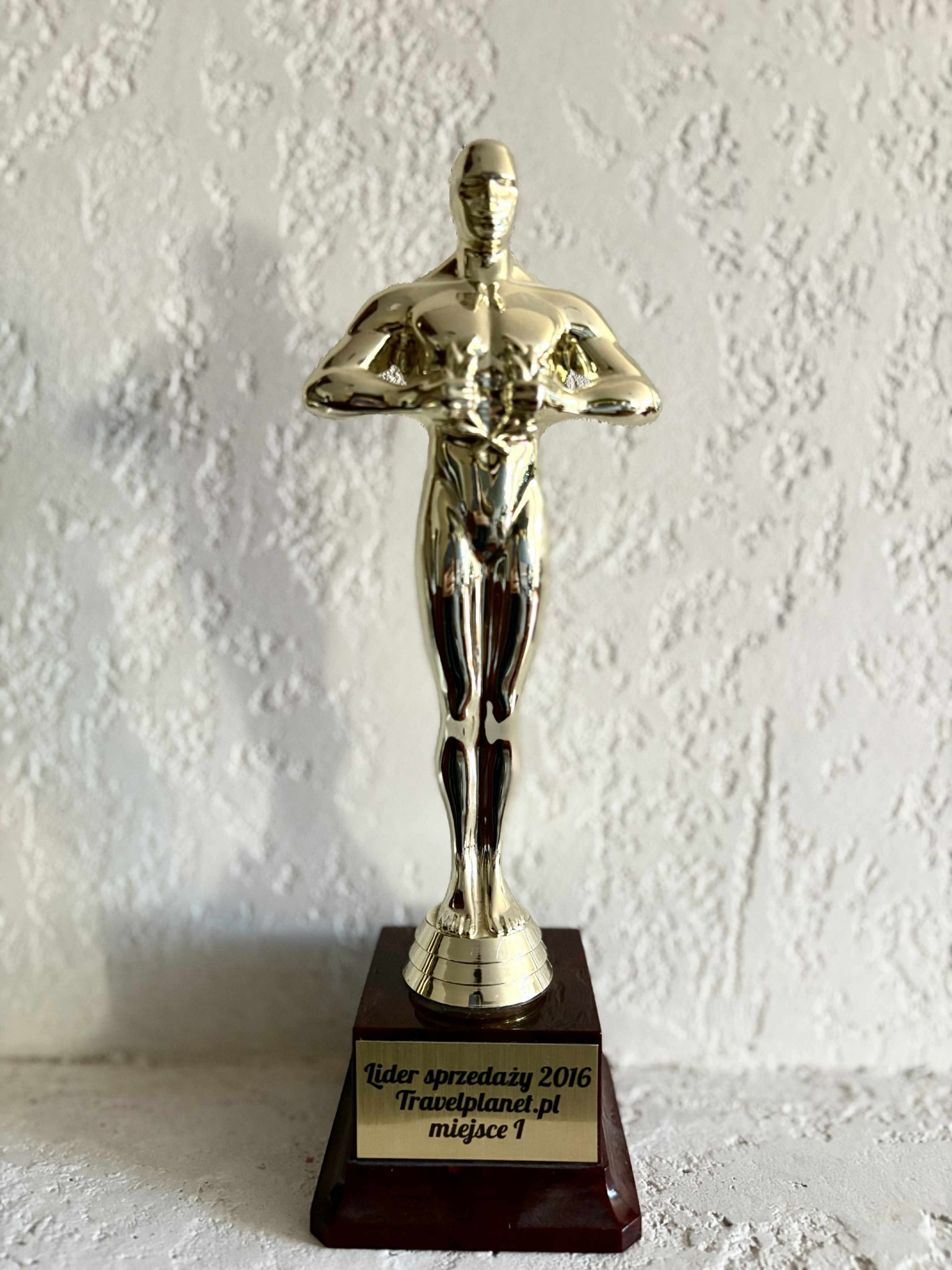 Złota statuetka 'Lider sprzedaży 2016 Travelplanet.pl miejsce 1' na tle białej, teksturowanej ściany. Nagroda za wyniki sprzedażowe w branży turystycznej.