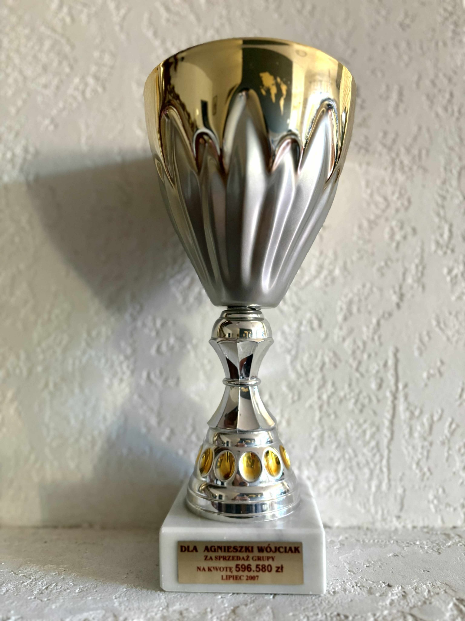 Błyszczący puchar w odcieniach złota i srebra, z wygrawerowaną dedykacją dla Agnieszki Wójciak za sprzedaż grupy na kwotę 596.580 zł, lipiec 2007, na tle teksturowanej ściany.