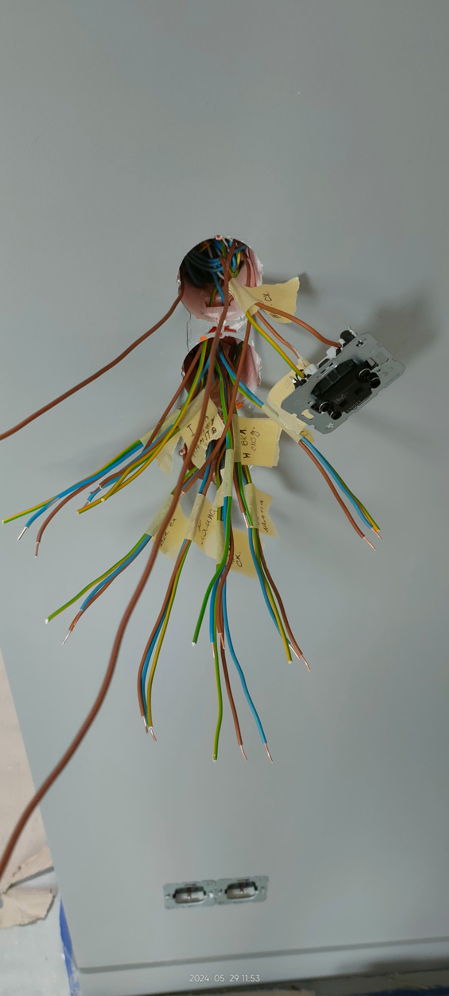 Przygotowane kable elektryczne z oznaczeniami, wychodzące ze ściany, przed montażem gniazdka. Widoczne przewody w różnych kolorach i fragment puszki elektrycznej.