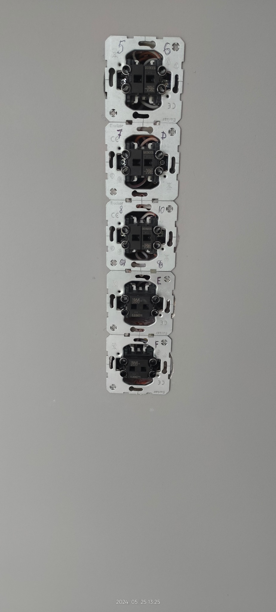 Rząd gniazdek elektrycznych Berker z numeracją, bez osłon, zamontowanych pionowo na szarej ścianie. Widoczne przewody i elementy montażowe.