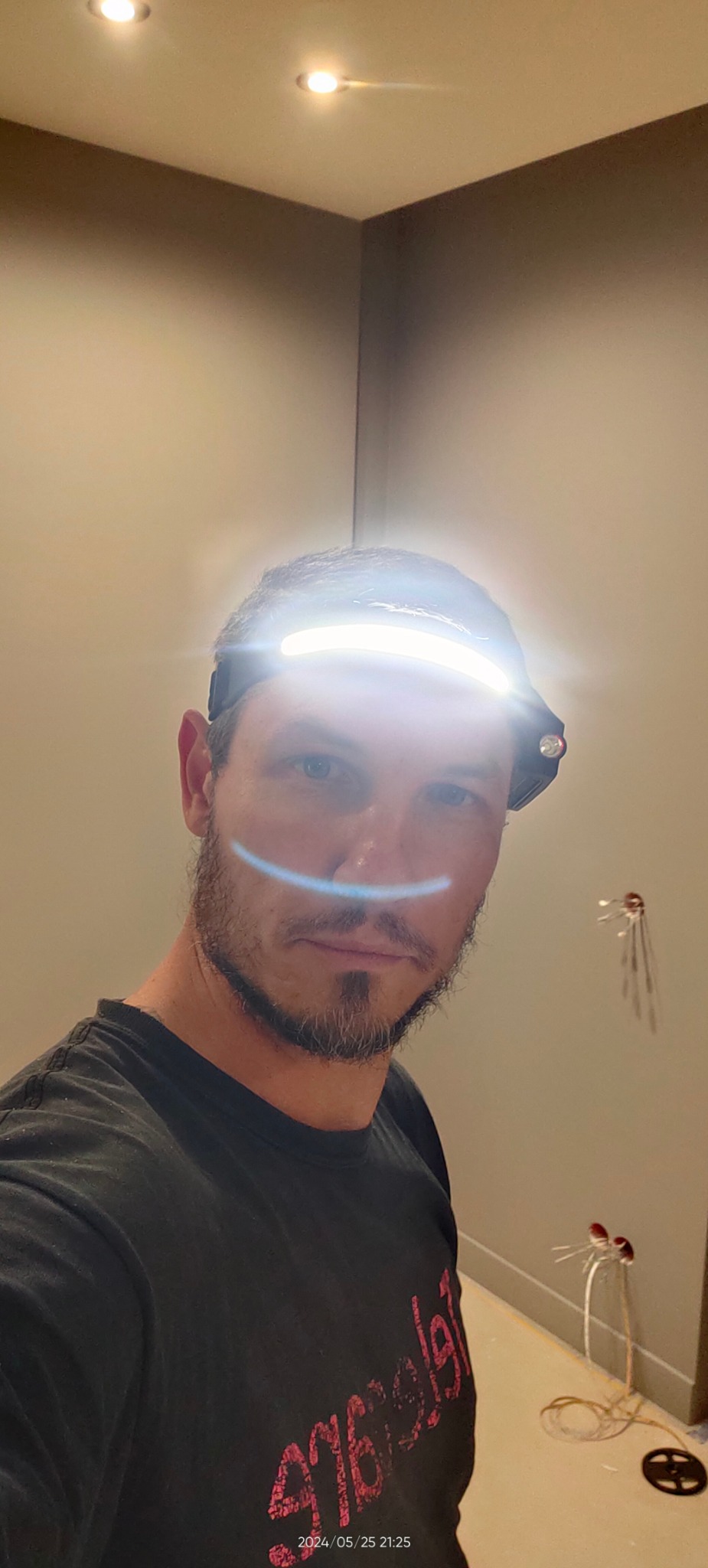 Elektryk w trakcie pracy, oświetlenie sufitowe zamontowane. Widoczne przewody w ścianie i lampka czołowa elektryka.