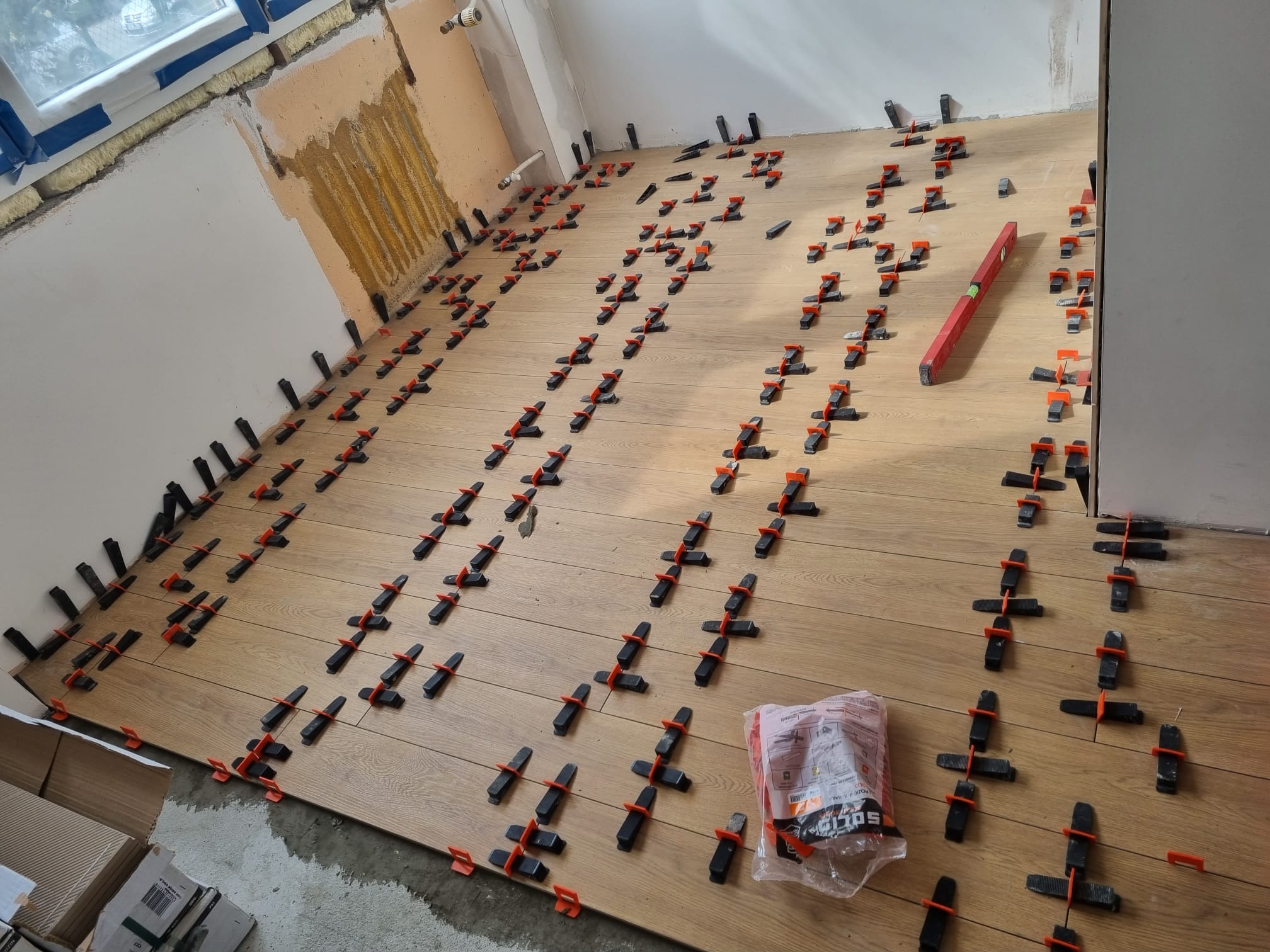 Układanie paneli podłogowych z użyciem klipsów poziomujących; widoczny poziomica i opakowanie klipsów.