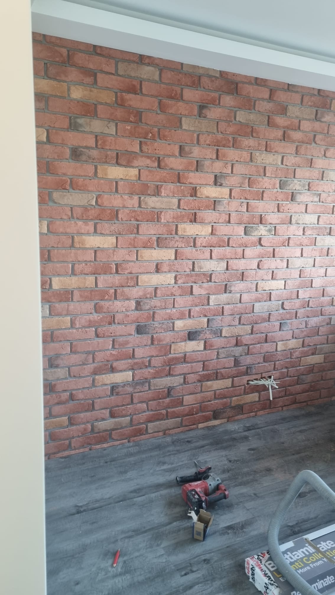 Ściana wyłożona dekoracyjną cegłą w odcieniach czerwieni i beżu, szara podłoga z narzędziami i materiałami budowlanymi, widoczne przewody elektryczne wystające ze ściany.