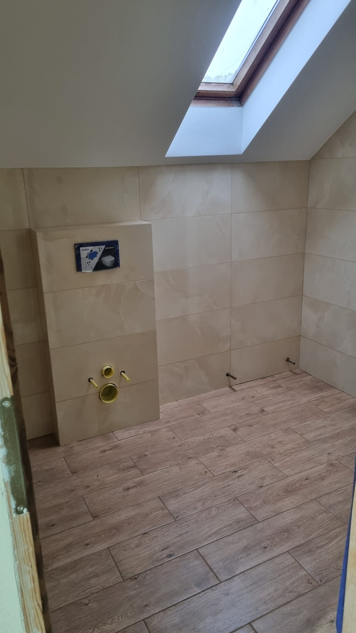 Wnętrze łazienki w trakcie remontu, widoczne beżowe płytki na ścianach i podłodze imitujące drewno, okno dachowe w skosie, oraz przygotowane przyłącza do armatury.