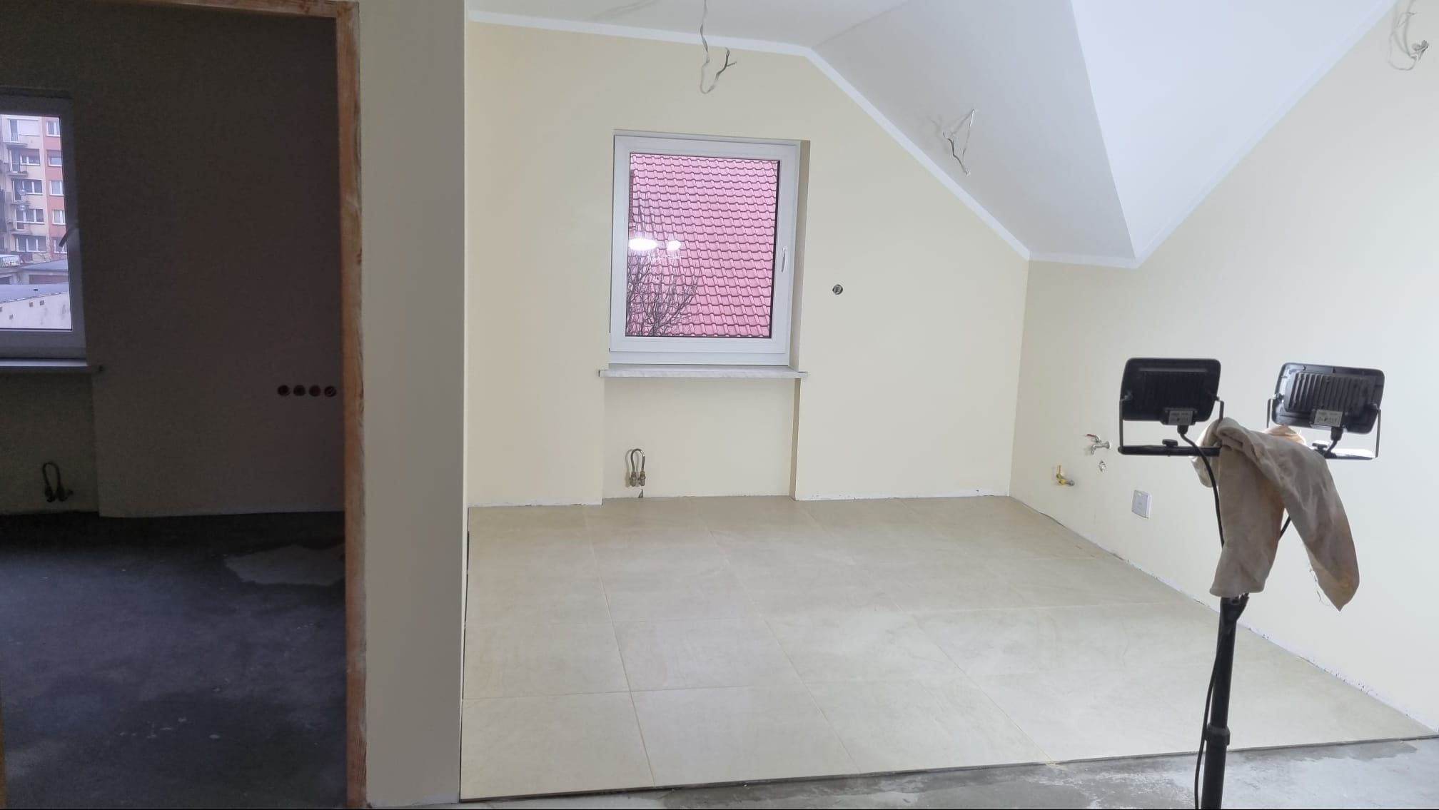 Pokój na poddaszu w trakcie remontu, z położoną jasną terakotą, świeżo pomalowanymi ścianami na kremowo, widoczne lampy budowlane i surowe wykończenie w przejściu do kolejnego pomieszczenia.