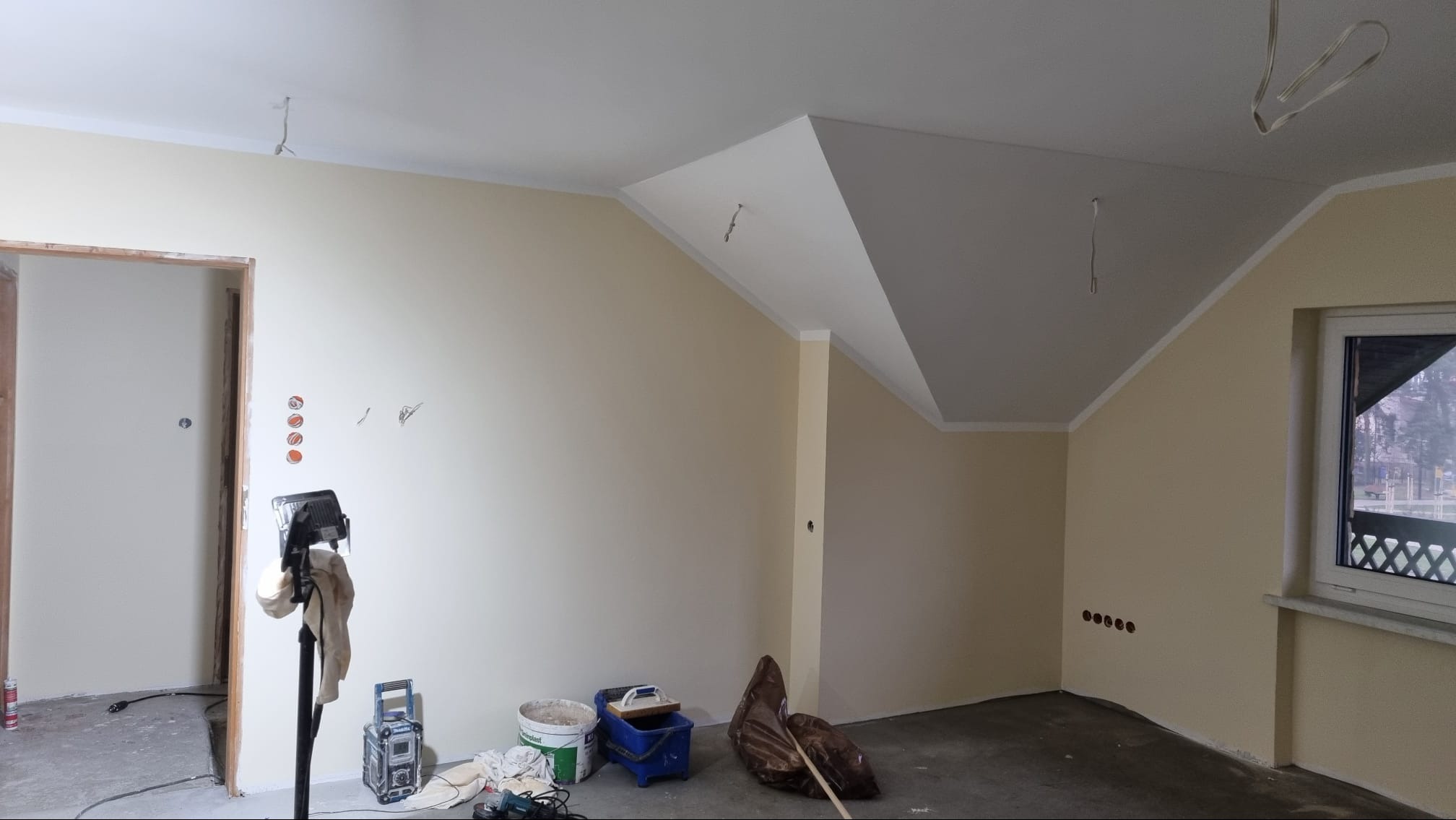 Wnętrze w trakcie prac wykończeniowych: świeżo pomalowane ściany na kremowo, widoczne puszki elektryczne, narzędzia malarskie, oświetlenie robocze i wiszące kable elektryczne.