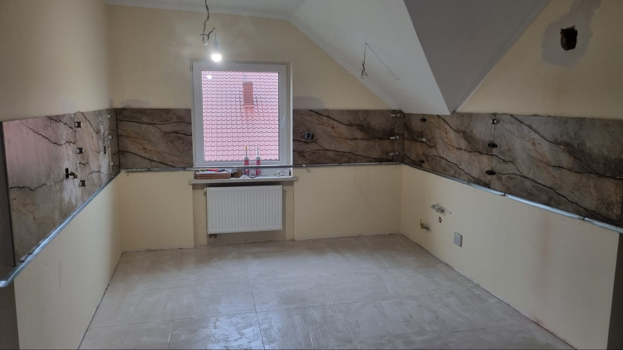 Poddasze w trakcie prac wykończeniowych: częściowo położone płytki imitujące marmur na ścianach, widoczne profile montażowe, grzejnik pod oknem, ściany w jasnym kolorze, surowe wykończenie podłogi.