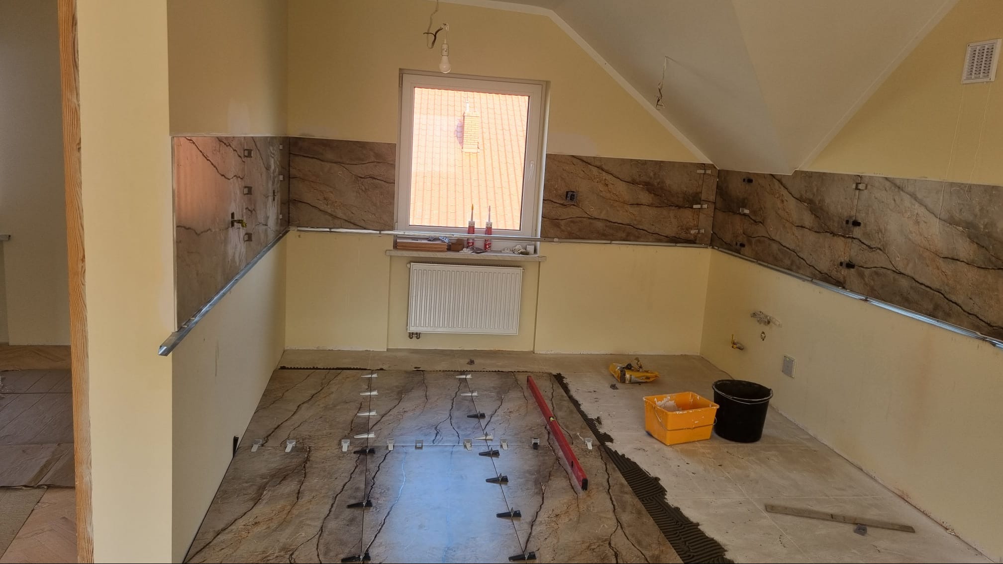 Układanie płytek imitujących marmur na podłodze i ścianach poddasza, widoczne poziomica, wiadra i krzyżyki dystansowe.