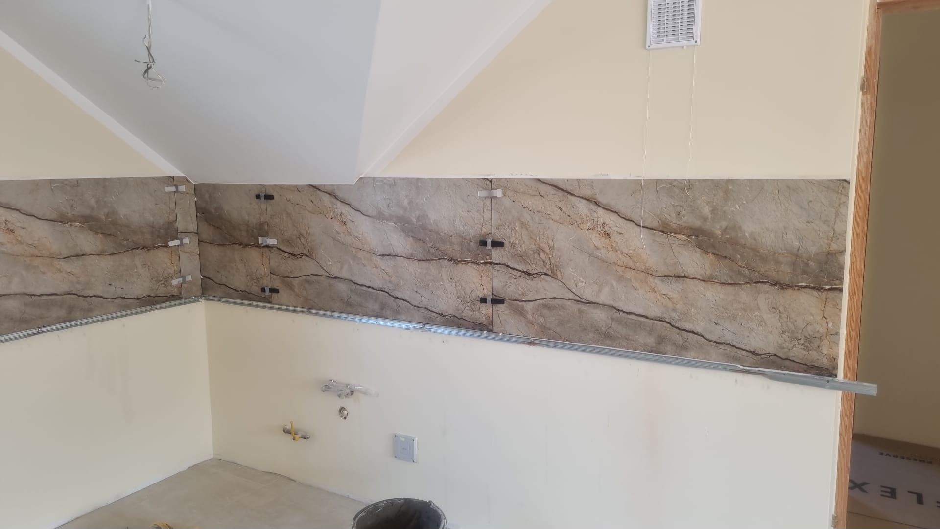 Ściana w trakcie układania płytek imitujących marmur, widoczne krzyżyki dystansowe i listwa montażowa, przygotowana do fugowania.
