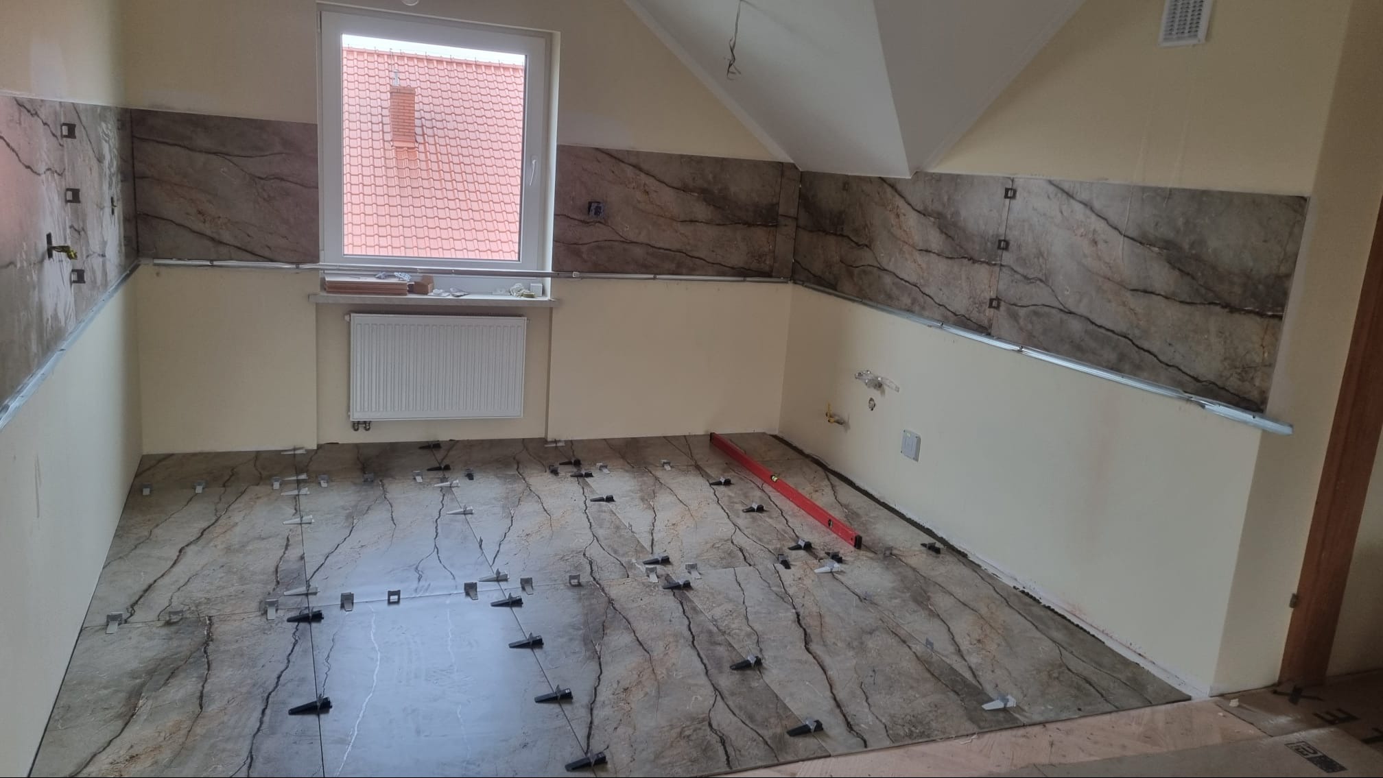 Montaż płytek imitujących marmur na podłodze i ścianach poddasza, widoczne systemy poziomowania płytek i poziomica.