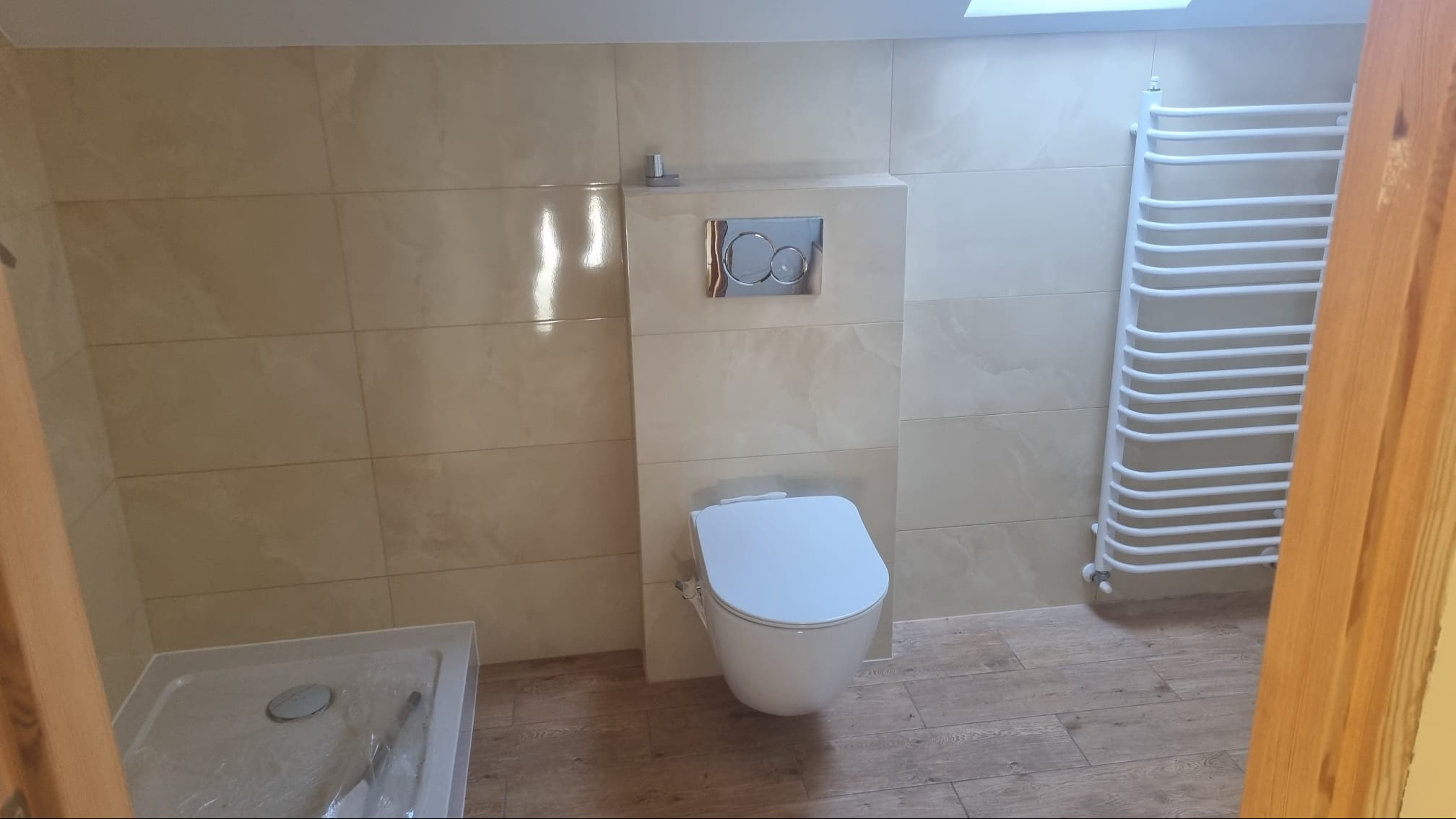 Nowoczesna łazienka w beżowych odcieniach z wiszącą toaletą, chromowanym przyciskiem spłukującym, grzejnikiem drabinkowym i częściowo widocznym brodzikiem.