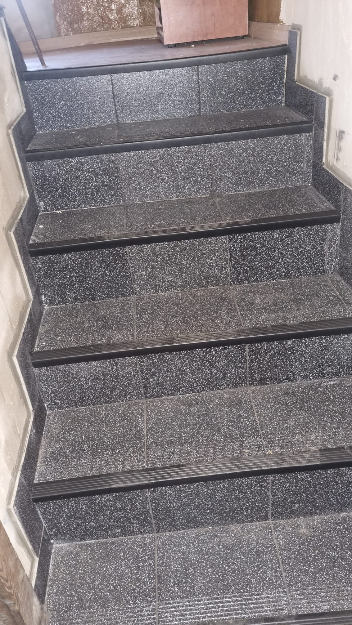 Schody wyłożone ciemną, szarą terakotą z czarnymi listwami antypoślizgowymi na krawędziach stopni, widoczne ślady zaprawy na ścianach.