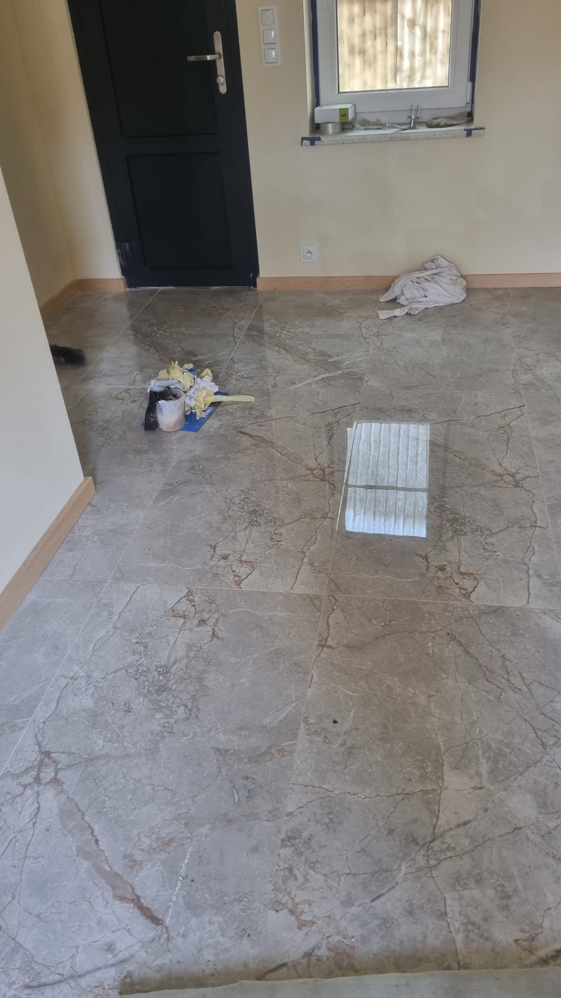 Nowa marmurkowa podłoga w pokoju, widoczne narzędzia glazurnicze i odbicie okna na błyszczącej powierzchni.