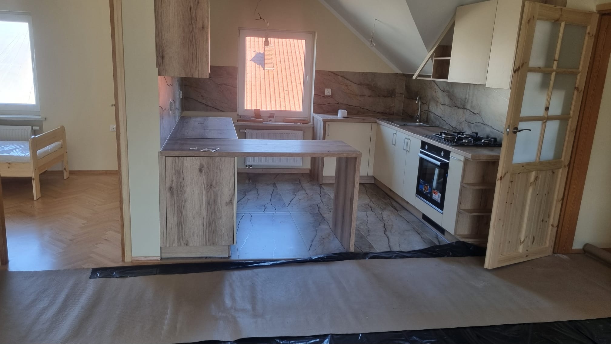 Jasna kuchnia w trakcie montażu, widoczne nowe szafki z drewnianymi frontami, płyta grzewcza, piekarnik, marmurowe płytki na ścianie, podłoga zabezpieczona papierem i folią ochronną, w tle widoczny...