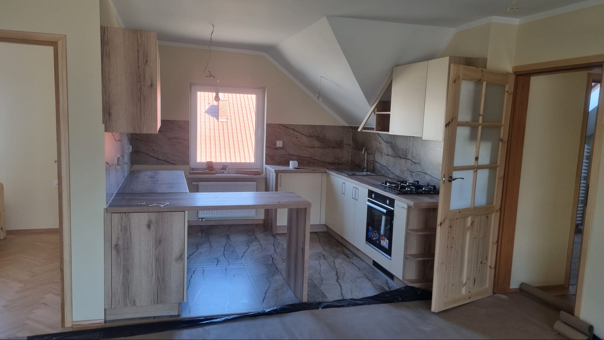 Nowoczesna kuchnia w trakcie wykańczania, z beżowymi szafkami, blatem imitującym marmur, wyspą kuchenną i drewnianymi drzwiami z szybkami, widok na okno dachowe.