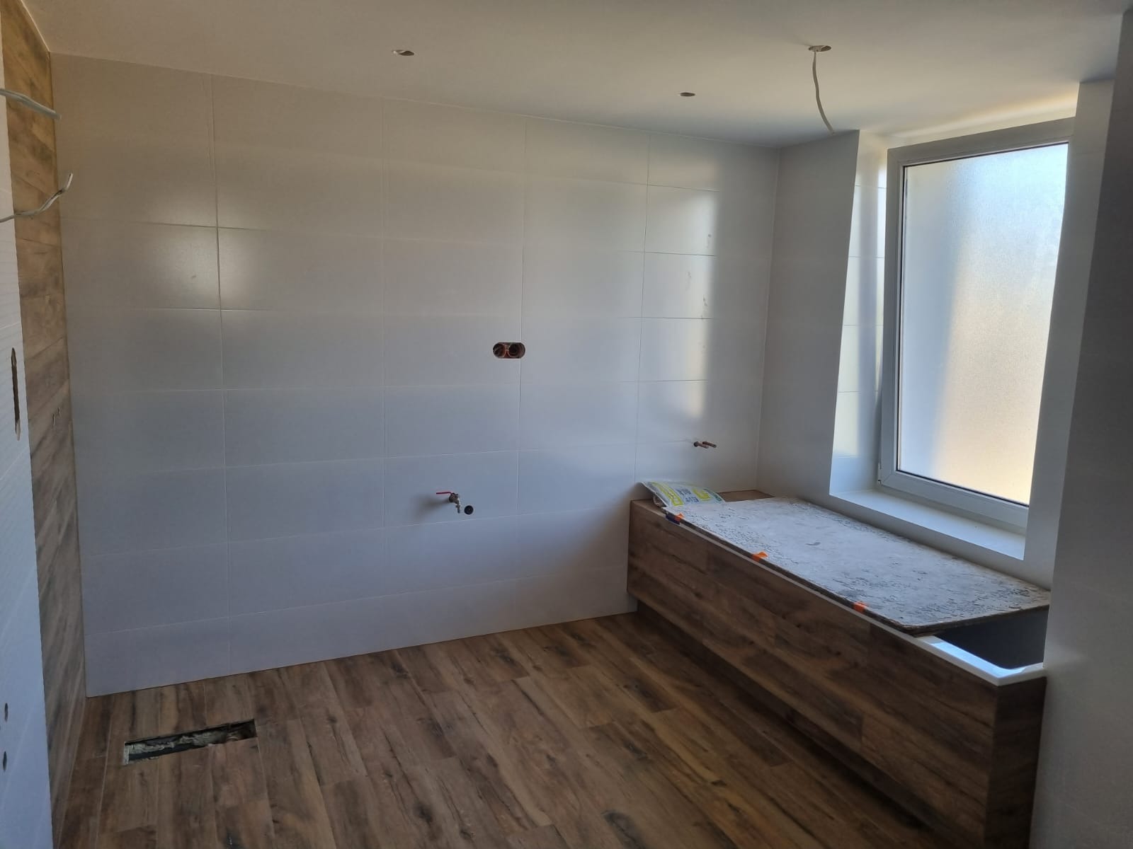 Wnętrze łazienki w trakcie prac wykończeniowych: ułożone białe płytki na ścianach, drewnopodobna podłoga, zabudowa wanny przy oknie, widoczne wystające kable i rury.