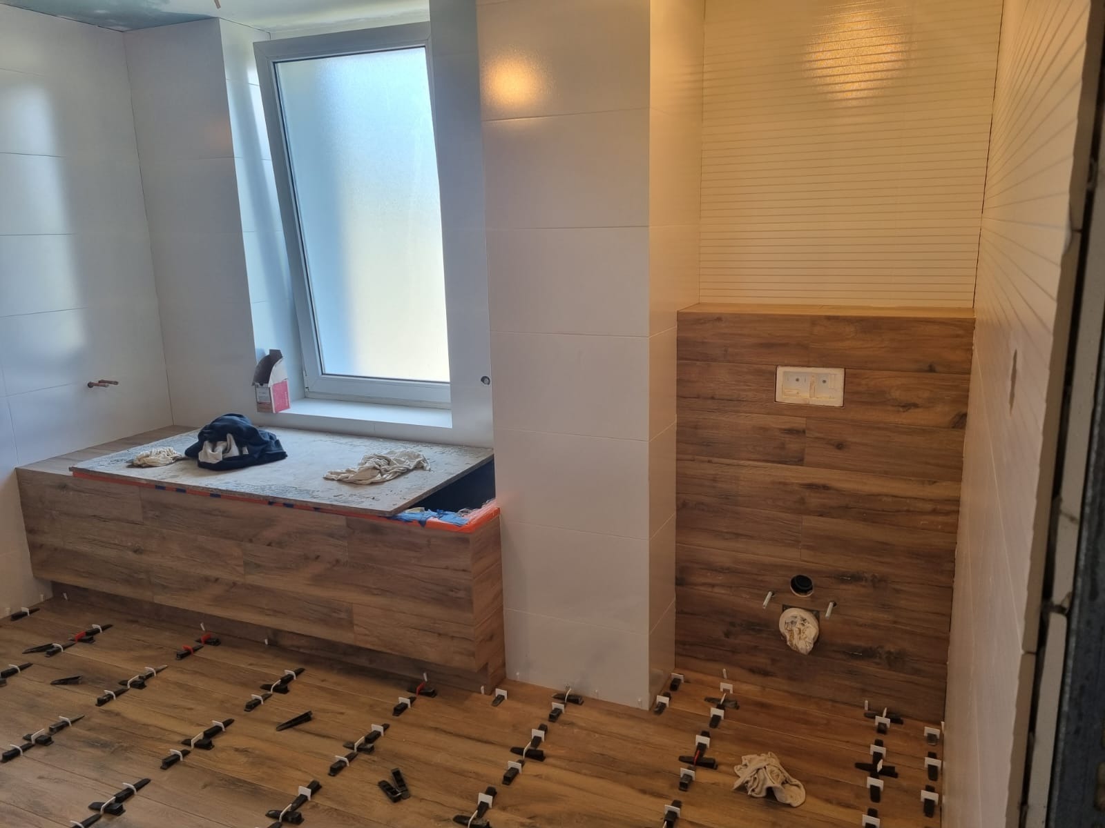 Łazienka w trakcie wykańczania: drewnopodobne płytki na podłodze i ścianach, białe płytki na ścianie z oknem, poziomowanie płytek za pomocą klipsów i klinów.