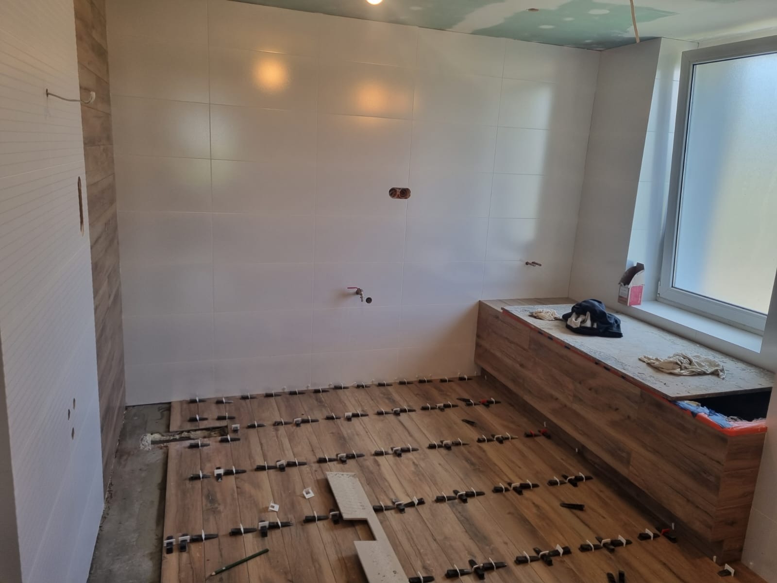 Łazienka w trakcie układania płytek podłogowych imitujących drewno, z białymi płytkami na ścianach i widocznymi elementami instalacji, takimi jak rury i gniazdka elektryczne.