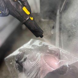 Autoart - Czyszczenie parowe elementu tapicerki samochodowej przez osobę w rękawicach, z użyciem parownicy z żółtymi akcentami, w pomieszczeniu z metalowymi elementami.