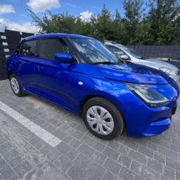 Błyszczący, granatowy samochód na kostce brukowej, z widocznym srebrnym autem w tle i błękitnym niebem z chmurami. Auto po detailingu, z wyraźnym odbiciem światła.